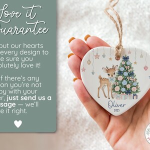 May include: Heart-shaped ceramic ornament featuring a fawn and Christmas tree design. The ornament has a string for hanging and includes the name "Oliver" and the year "2025". The ornament is held in a hand.