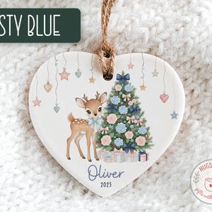 May include: Heart-shaped Christmas ornament with a deer and Christmas tree design. The ornament features a deer with a blue bow, a decorated tree, and the name "Oliver" with the year "2025". The text "Dusty Blue" is at the top.