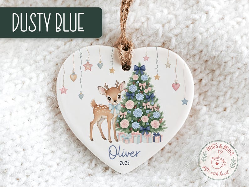 May include: Heart-shaped Christmas ornament with a deer and Christmas tree design. The ornament features a deer with a blue bow, a decorated tree, and the name "Oliver" with the year "2025". The text "Dusty Blue" is at the top.