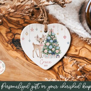 May include: Heart-shaped ceramic Christmas ornament featuring a deer and a decorated tree. The ornament has a jute string for hanging and includes the name "Grace" and the year "2025". Pastel stars and hearts decorate the top.