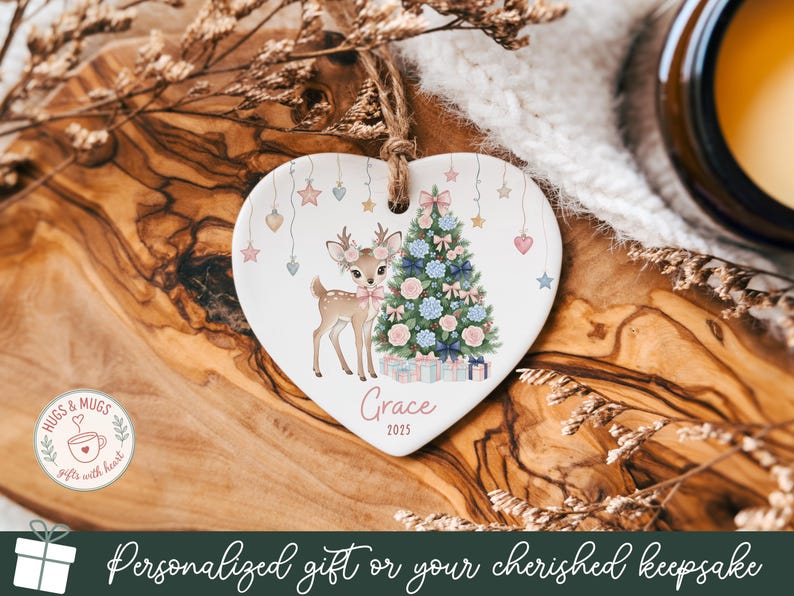 May include: Heart-shaped ceramic Christmas ornament featuring a deer and a decorated tree. The ornament has a jute string for hanging and includes the name "Grace" and the year "2025". Pastel stars and hearts decorate the top.