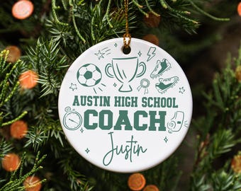 May include: White ceramic ornament with green sports-themed illustrations and text. The ornament says "Austin High School Coach Justin" with images of a soccer ball, trophy, and cleats. A gold string is attached for hanging.