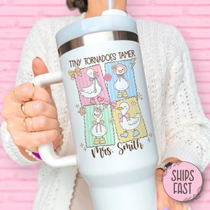 May include: White insulated tumbler with a handle, featuring a design with four cartoon ducks in pastel-colored squares. The text "TINY TORNADOES TAMER Mrs. Smith" is printed on the tumbler. The tumbler has a light blue lid and a white handle.