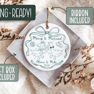 May include: White ceramic wedding ornament with a green illustration of two geese toasting with champagne glasses. The ornament reads "Merry & Married", "Olivia & Mike 2025", and includes a gift box and ribbon. The ornament is ready for gifting.