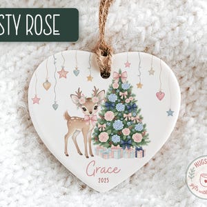 May include: Heart-shaped Christmas ornament with a deer and Christmas tree design. The ornament features a deer with floral antlers and a pink bow, a decorated tree, and the name "Grace" with the year 2025. The text "Dusty Rose" is at the top.