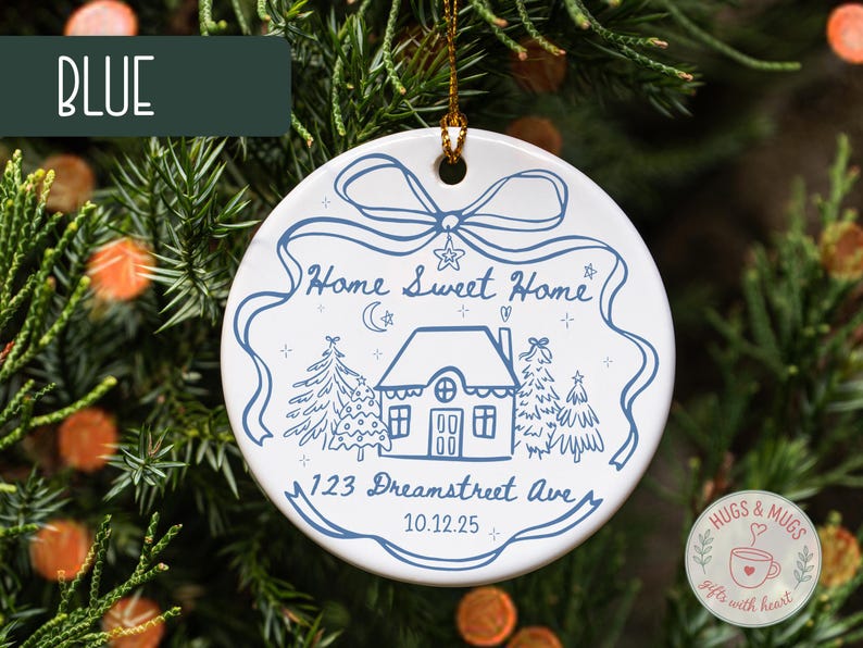 May include: White ceramic Christmas ornament with a blue illustration of a house, trees, and the text "Home Sweet Home" and "123 Dreamstreet Ave 10.12.25". The ornament is hanging on a Christmas tree.