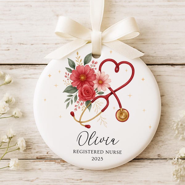 Puede incluir: Adorno redondo blanco con una cinta crema, con un diseño floral y un estetoscopio rojo. El adorno incluye el nombre "Olivia" y el texto "Registered Nurse 2025".