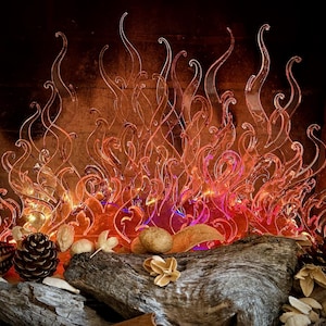 May include: A decorative fireplace insert featuring translucent, flame-shaped acrylic cutouts in shades of orange and red. The structure is illuminated from below, creating a glowing effect, and is arranged behind natural wood, pinecones, and dried botanicals.