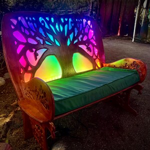 Puede incluir: Banco de madera con un diseño de árbol de la vida tallado en el respaldo. El banco está iluminado con un arcoíris de luces LED. Tiene un cojín verde y está hecho de madera.