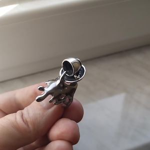 May include: A silver pendant with an abstract design, resembling a liquid splash. The pendant hangs from a small, circular bail. The jewellery is held in a person's hand.