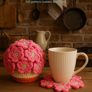 Könnte beinhalten: Ein gehäkeltes Blumentopf- und Untersetzer-Set. Die Topfabdeckung ist rosa mit grünen Zentren, und der Untersetzer hat eine passende Blumenform. Eine weiße Tasse steht auf dem Untersetzer. Der Text lautet "Flower pot Coaster Set" und "Pdf pattern instant download."