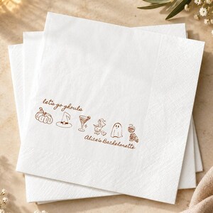 May include: A stack of square white paper napkins featuring a brown line art design. The print includes the text "let's go ghouls" and "Alice's Bachelorette" alongside small illustrations of a pumpkin, witch hat, cocktail, ghost, and skull.