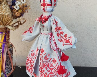 Puede incluir: Muñeca hecha a mano con un vestido bordado tradicional. El vestido gris claro presenta patrones florales y geométricos rojos, mangas largas y un tocado a juego. Un collar de cuentas rojas y un cinturón con borlas completan el atuendo.