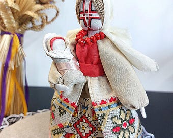 Puede incluir: Muñeca de tela hecha a mano con diseño tradicional. La muñeca lleva un top rojo, un velo blanco y una falda con bordados coloridos. Sostiene una muñeca más pequeña envuelta en tela blanca. Un collar de cuentas rojas adorna el cuello de la muñeca.