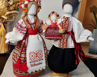Puede incluir: Dos muñecas ucranianas hechas a mano, vestidas con ropa bordada tradicional. La muñeca femenina lleva un vestido rojo y blanco con un tocado floral. La muñeca masculina lleva una camisa blanca, pantalones negros y un sombrero de piel. El hombre sostiene una muñeca más pequeña.