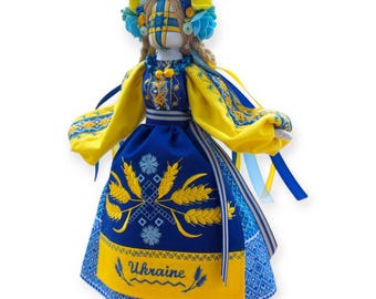 May include: A handmade doll in traditional Ukrainian attire. The doll features a blue and yellow dress with wheat embroidery and the word "Ukraine". It has a floral headdress and long, flowing ribbons. The doll's face is painted with a traditional design.