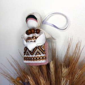 May include: A handmade fabric doll with a white face, a patterned dress in brown, white, and pink, and a head covering. The doll has wooden bead accents and a lavender ribbon for hanging. The doll is displayed with a bundle of wheat.