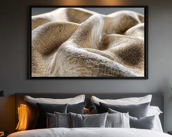 May include: A framed canvas print featuring a close-up of textured, beige fabric with flowing, curved lines. The artwork is displayed above a bed with gray pillows and a snake plant in a black pot.
