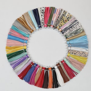 Pode incluir: Um arranjo circular de inúmeras borlas de couro sintético numa grande variedade de cores sólidas e padrões, incluindo estampados de leopardo, florais e riscas. Cada borla apresenta uma tampa de metal prateado com um pequeno anel.