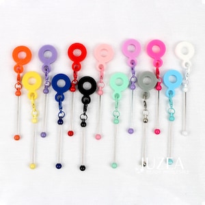 Può includere: Una collezione di ciondoli decorativi colorati con una parte superiore circolare e una catena pendente. I ciondoli sono disponibili in vari colori, tra cui arancione, viola, rosso, rosa, verde, grigio e blu. Ogni ciondolo ha una piccola perla alla fine della catena pendente.