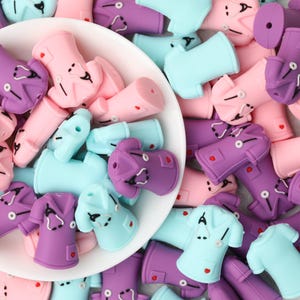 May include: A collection of silicone beads shaped like medical scrub tops in shades of light blue, pink, and purple. Each bead features a molded stethoscope detail, a small pocket, and a tiny red heart accent, with a hole for threading.