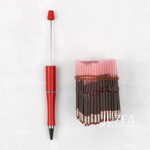 May include: A red pen with a silver tip and a bundle of red and clear glass beads. The beads are held together with a brown rubber band. The pen is next to the beads. The text "ZEA BEADS WHOLESALE" is visible.