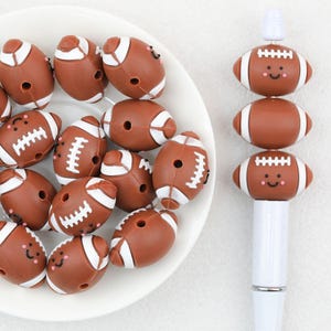 May include: A collection of brown silicone football beads with white laces and smiling faces, some loose in a white bowl and others stacked vertically on a white pen. Each bead features a central hole for threading, showcasing a smooth texture.