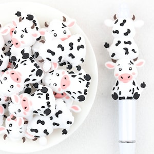May include: A collection of small, white and black cow-shaped silicone charms with pink snouts and ears, piled together on a white surface, alongside a white pen topped with two stacked cow figures featuring the same black and white pattern.