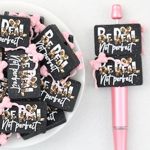 Op de afbeelding: Een verzameling rechthoekige zwarte kralen met de tekst "BE REAL Not perfect" in witte en luipaardprint letters, geaccentueerd met kleine roze strikjes. Eén kraal zit vast aan een roze pen, terwijl andere los in een witte kom liggen.