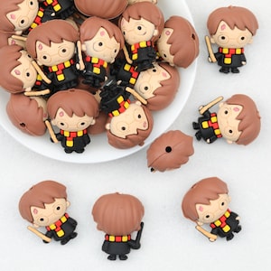 May include: A collection of small, three-dimensional silicone beads shaped like a cartoon character with brown hair, round glasses, a lightning bolt scar, and a black robe with a red and yellow striped scarf, holding a small wand in one hand.