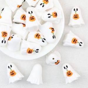 Könnte beinhalten: Eine Sammlung weißer geisterförmiger Perlen, die jeweils einen kleinen orangefarbenen Jack-o'-Lantern-Kürbis in der Mitte halten. Die Geister haben schwarze Augen und Münder, wobei einige Perlen auf einem weißen Teller und andere verstreut liegen.