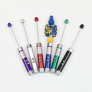May include: A collection of laser pointers in various colors: purple, silver, red, blue, green, and black. Each pointer has a silver body, a button, and a matching colored tip. A charm with the words "BE KIND" and "DOWN SYNDROME" is also present.