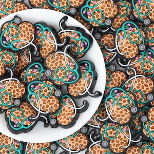 May include: A collection of flat, heart-shaped charms featuring a tan and brown leopard print pattern. Each charm is outlined in black and decorated with a teal stethoscope graphic that includes small pink flowers along the tubing.