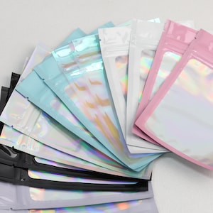May include: A collection of flat, rectangular resealable pouches arranged in a fan shape. The pouches feature a clear, iridescent window on the front and come in various solid colors including black, light blue, white, and soft pink.