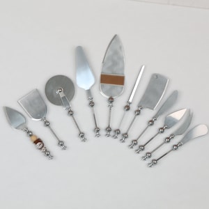 May include: A set of ten silver-toned metal cheese serving utensils arranged in a fan shape. Each tool features a slender handle with decorative spherical accents and a unique blade shape, including a pizza cutter, cleaver, and various spreaders.