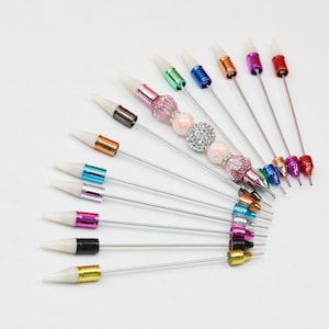 May include: A selection of decorative pins with white tips and colourful metallic accents. The pins feature various bead designs, including clear and pink rhinestones, arranged in a fan shape against a white background.