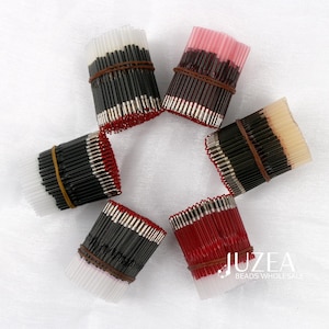 Op de afbeelding: Zes bundels kralen, elk vastgezet met een bruine elastiek. De kralen zijn zwart, rood en wit, met zilverkleurige uiteinden. De afbeelding is goed belicht, waardoor de kleuren en texturen goed zichtbaar zijn.