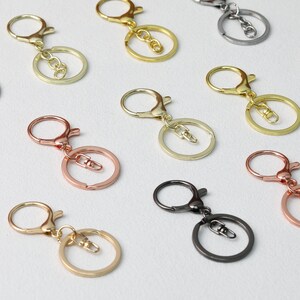 Pode incluir: Variedade de chaveiros em diversos acabamentos metálicos, incluindo dourado, ouro rosa, prateado e preto. Cada chaveiro possui um mosquetão e um aro bipartido para prender chaves ou pingentes. Em um fundo branco.