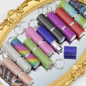 Könnte beinhalten: Eine Sammlung von Strass-Lippenstifthaltern in verschiedenen Farben, darunter Rosa, Grün und Blau, mit silbernen Schlüsselanhängern. Die Halter sind auf einer reflektierenden Oberfläche in einem goldfarbenen Dekotablett angeordnet. Ein kleiner, blau verzierter Würfel ist ebenfalls vorhanden.