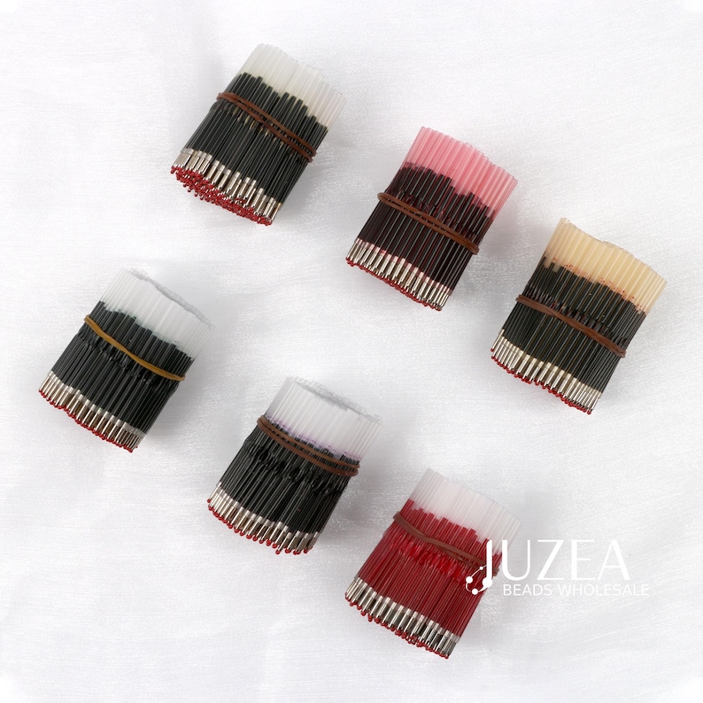May include: Bundles of colorful craft pins, each secured with a brown rubber band. The pins display a variety of colors, including black, white, pink, and red. The tips of the pins are silver. The image is taken from above, with a white background.