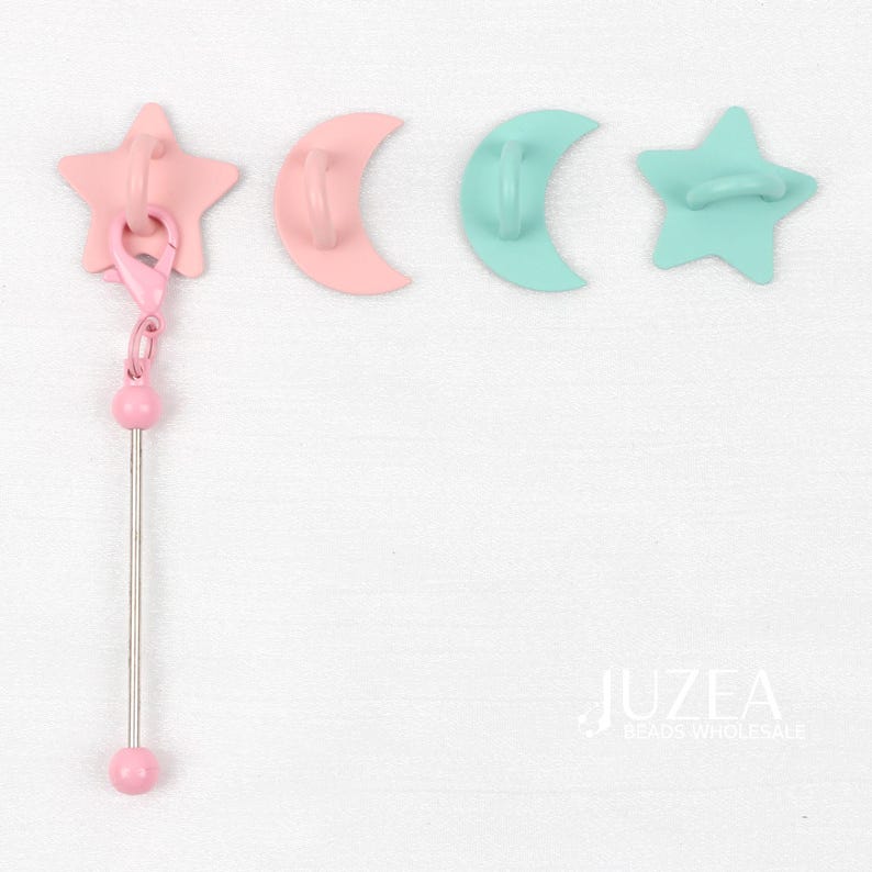 Puede incluir: Una colecci&oacute;n de colgantes en forma de estrella y luna en colores pastel. Un colgante de estrella rosa est&aacute; conectado a una cadena de cuentas rosa con una varilla plateada. Los otros colgantes son una luna creciente rosa, una luna creciente turquesa y una estrella turquesa.