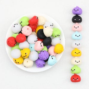 Puede incluir: Una colección de pequeñas cuentas redondas con forma de bola de masa en varios colores, incluyendo rojo, amarillo, verde, azul, morado, negro, blanco y rosa. Cada cuenta presenta una cara sonriente impresa con dos ojos y una boca.