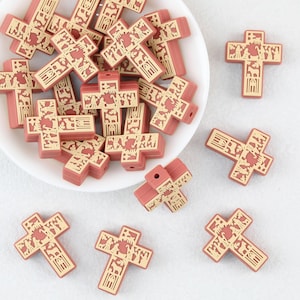May include: A collection of cross-shaped beads in a muted terracotta red color. Each bead features a recessed, light beige graphic design depicting a nativity scene with figures and animals. The beads have a central hole for threading purposes.