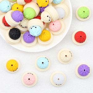 May include: A collection of small, round, multi-coloured silicone beads shaped like steamed buns. Each bead features a smiling face, a central hole, and a cream-coloured base. The assortment includes red, yellow, blue, purple, green, and pink colours.