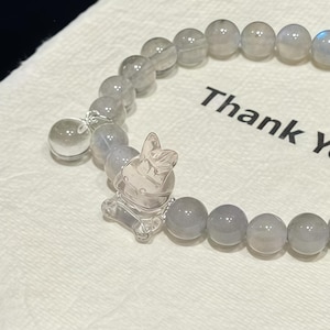 May include: A bracelet featuring grey, spherical beads and a clear bunny charm. The bracelet is displayed on a white cloth, with the words "Thank You" printed on it.