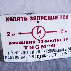 May include: A rectangular white metal sign with red text and symbols. The sign features the text "КОПАТЬ ЗАПРЕЩАЕТСЯ" and "ОХРАННАЯ ЗОНЯ КАБЕЛЯ". A lightning bolt symbol is in the center, with arrows and the number "2 m" on either side. The sign also includes additional text.
