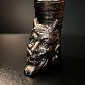 May include: A silver-coloured shot glass featuring a devil's head. The devil's face is intricately detailed with horns, a moustache, and a goatee. The glass has a cylindrical shape with a decorative rim.