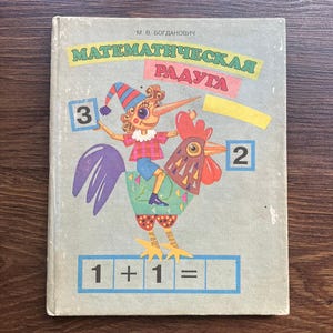 Op de afbeelding: Een lichtgrijs wiskundeboek voor kinderen met de titel M. V. Bogdanovich MATEMATICHESKAYA RADUGA. De kaft bevat een kleurrijke illustratie van een figuur met een gestreepte hoed op een haan, met de cijfers 3 en 2 en de vergelijking 1 + 1 =.