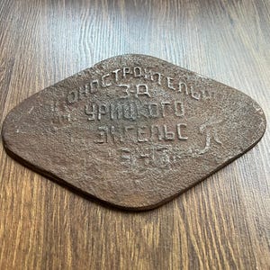 Puede incluir: Una placa de metal oxidado en forma de diamante con una superficie texturizada. La placa presenta texto en cirílico en relieve que dice "КОНОСТРОИТЕЛЬНЫЙ З-Д ИМ. УРИЦКОГО Г. ЭНГЕЛЬС". El metal tiene un aspecto marrón oscuro y desgastado con oxidación visible.