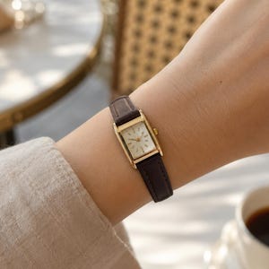 May include: A rectangular wristwatch featuring a gold-tone case and a white dial with gold-tone hour markers and hands. The watch is secured with a slim, dark brown leather strap that has visible stitching along the edges and a metal buckle.